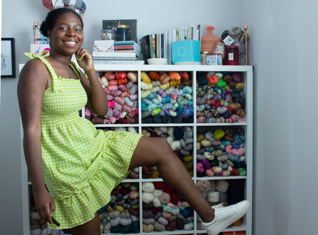 Black woman with one leg up in lime green dress. Standing in front of a storage unit filled with yarn